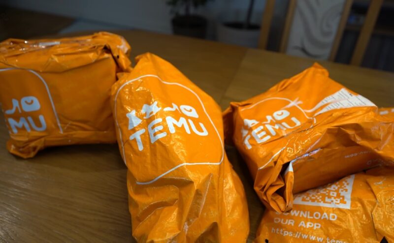 Tri narandžasta paketa sa logotipom Temu postavljena na drvenom stolu, spremna za isporuku kupcima